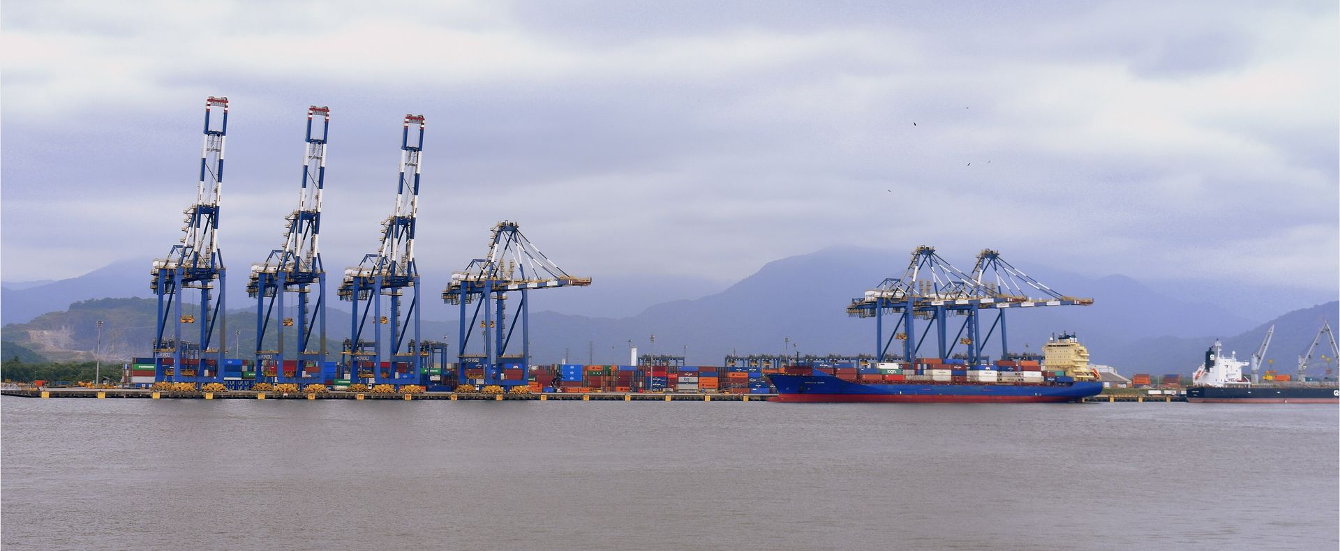 Terminal de contêineres do Porto de Santos, com guindastes e um navio porta-contêineres atracado, serra ao fundo